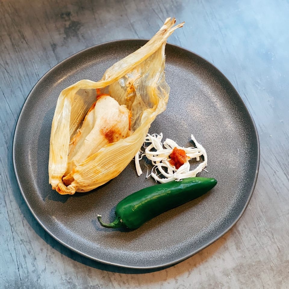 A tamale unwrapped on a dark plate with shredded cheese, salsa, and a jalapeño.